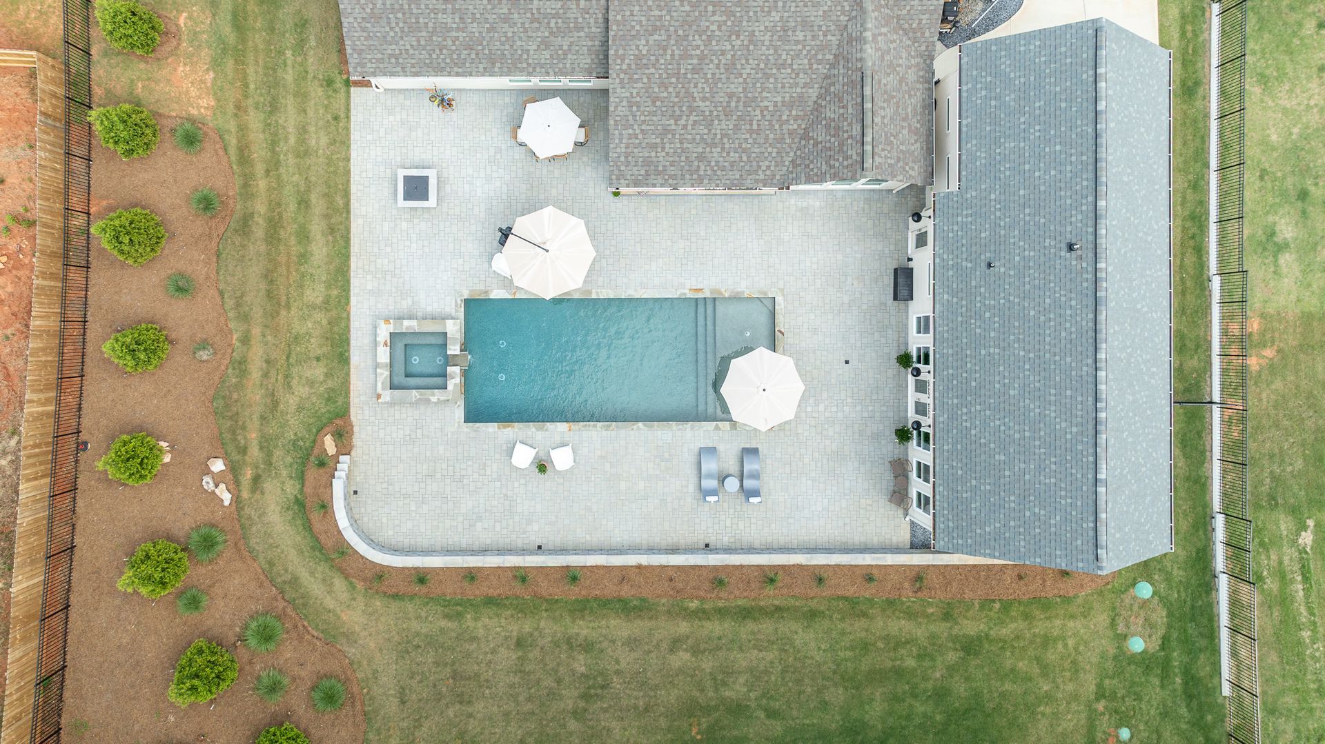 Aerial view of a backyard patio with table, umbrellas, a house, and surrounding grass and garden rows.