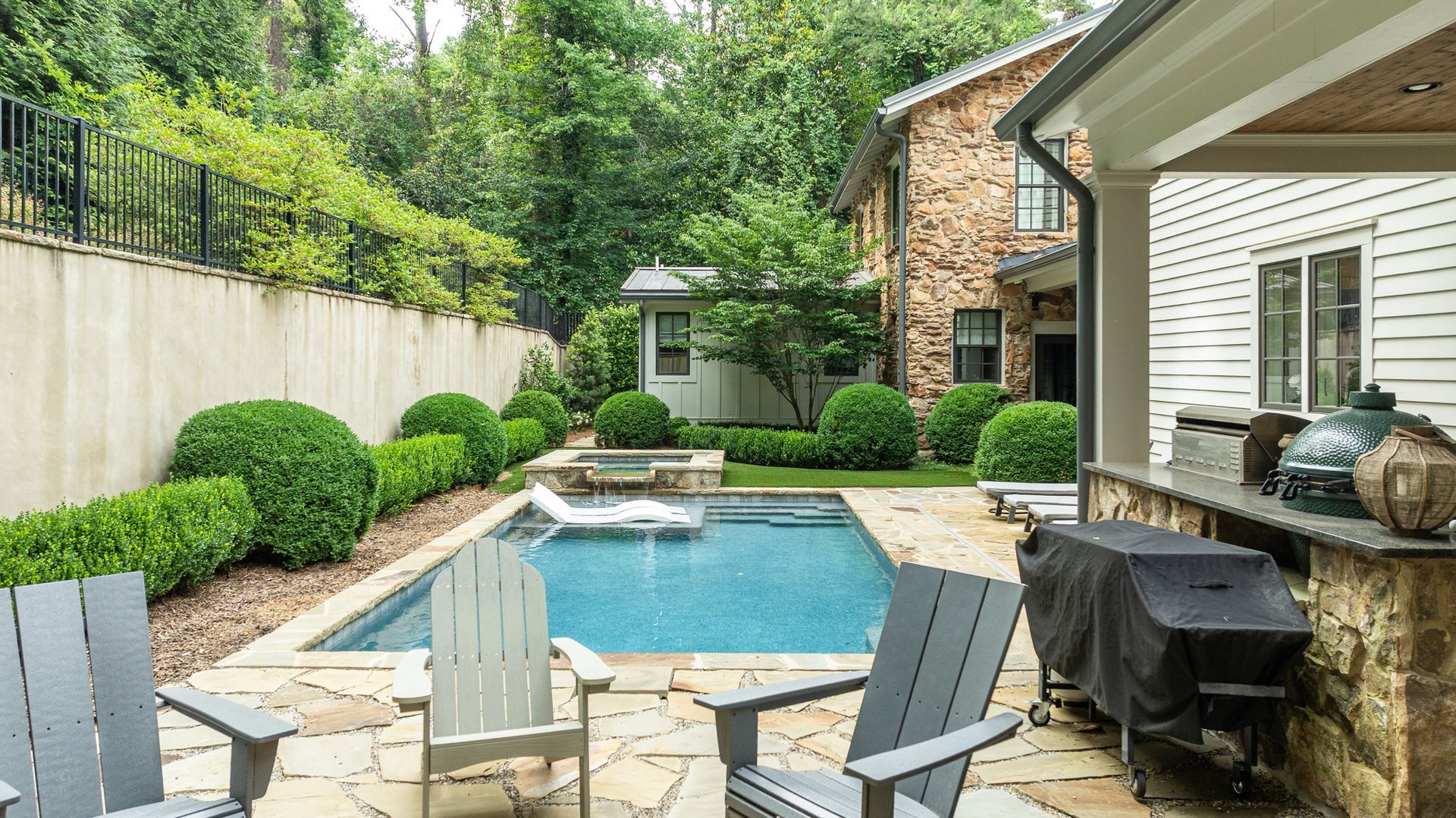 Backyard patio with a blue pool, stone seating area, grill, and white house beside a brick home.