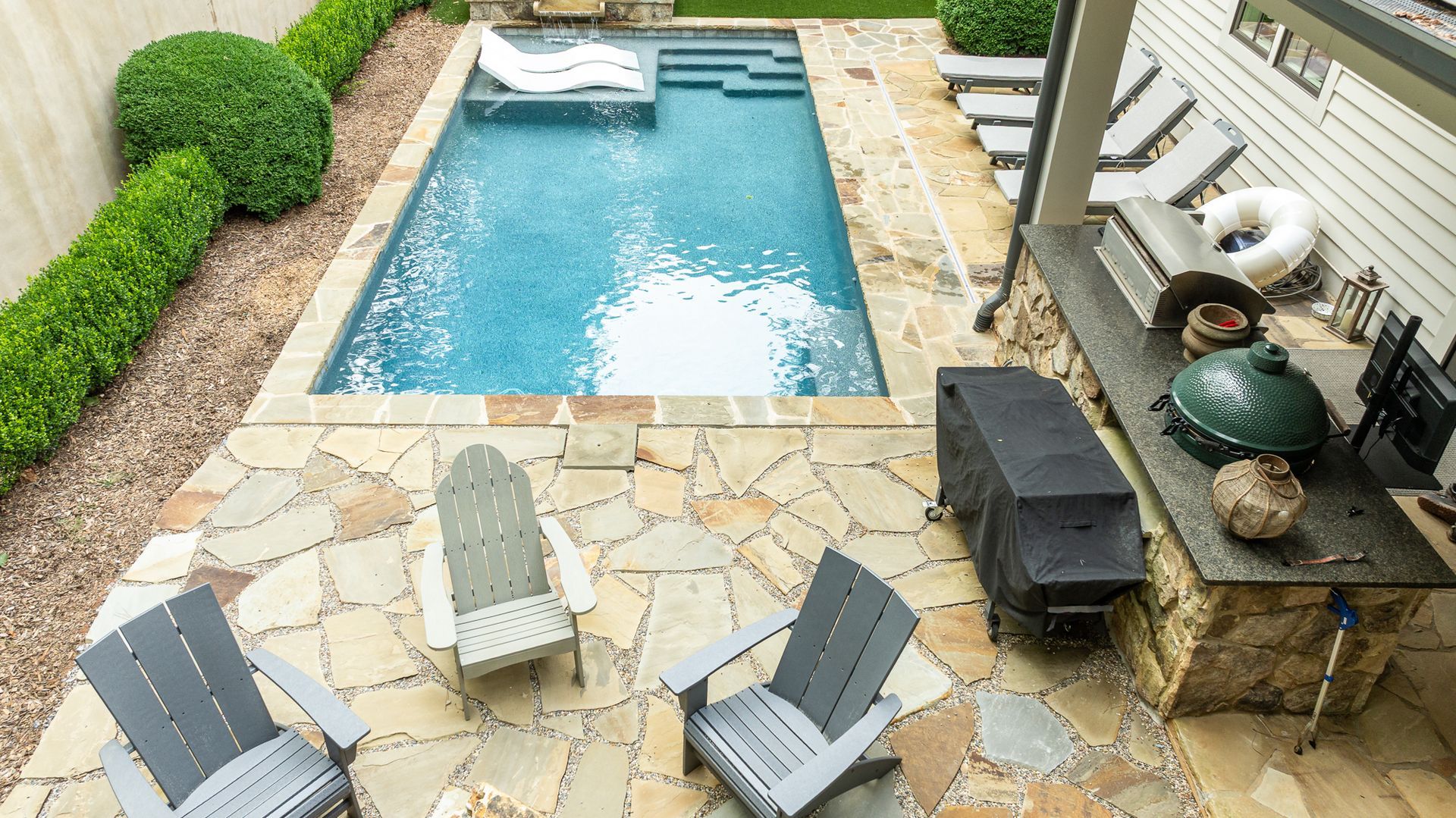 Backyard pool with stone patio, lounge chairs, grill, and green hedges