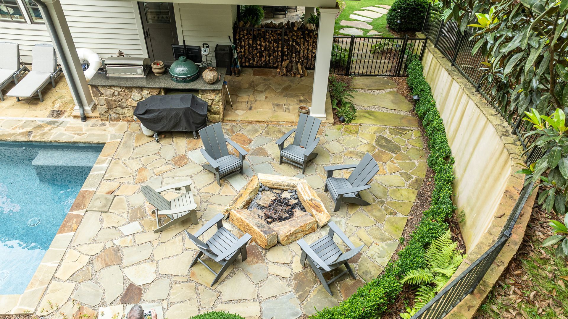 Backyard patio with stone fire pit, gray chairs, pool edge, and greenery along a curved wall