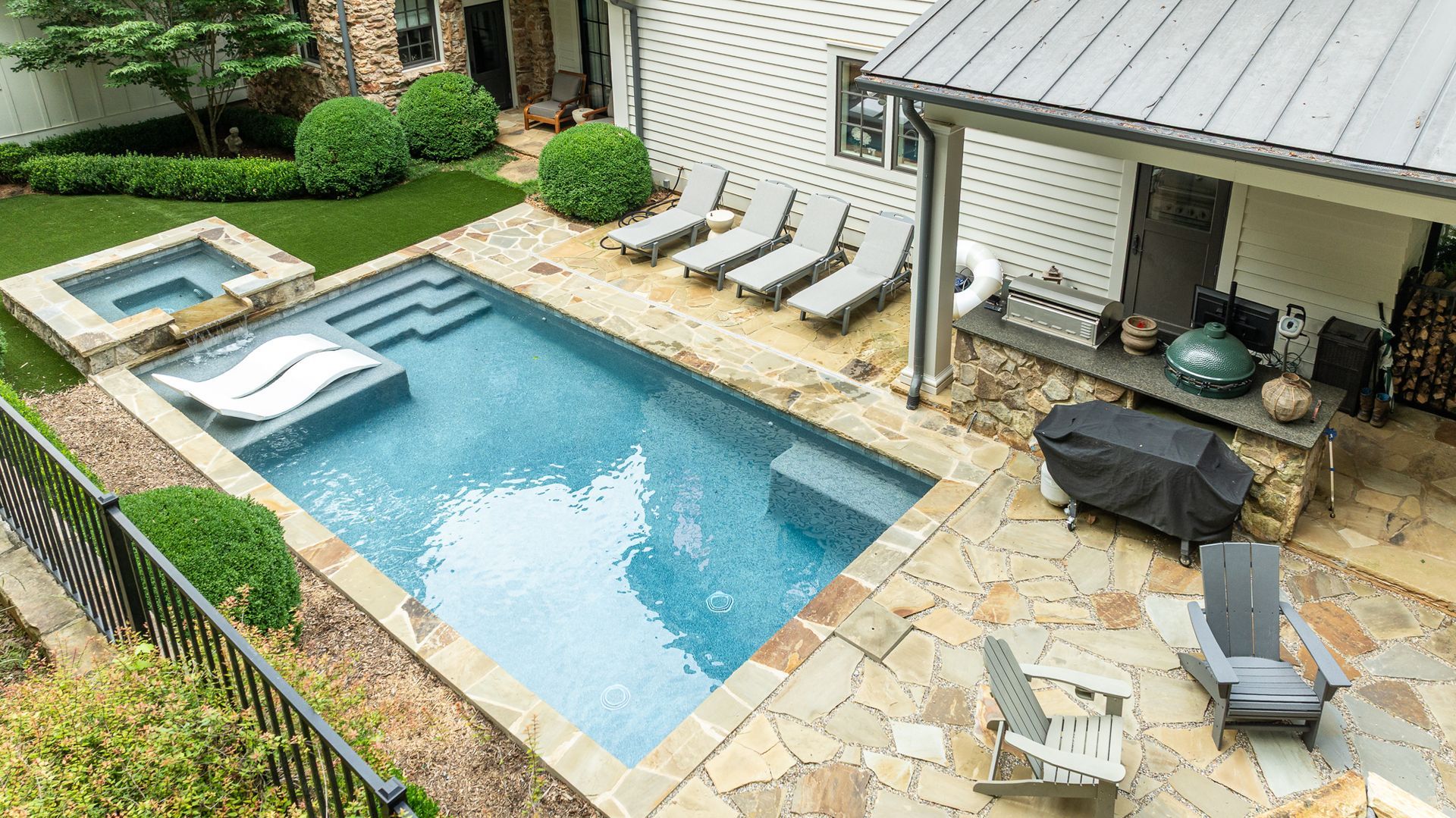 Backyard pool with stone patio, hot tub, lounge chairs, and covered grill area