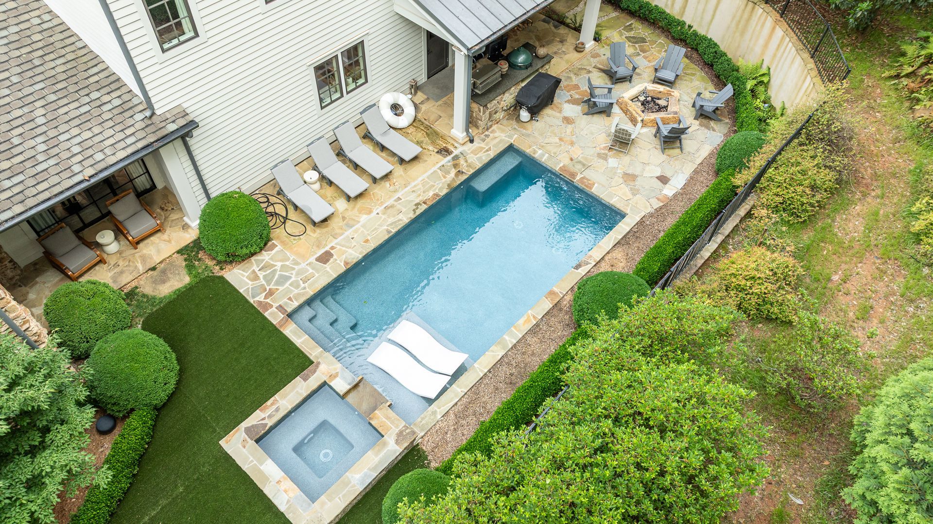 Aerial view of a backyard with a pool, hot tub, lounge chairs, and patio seating next to a house