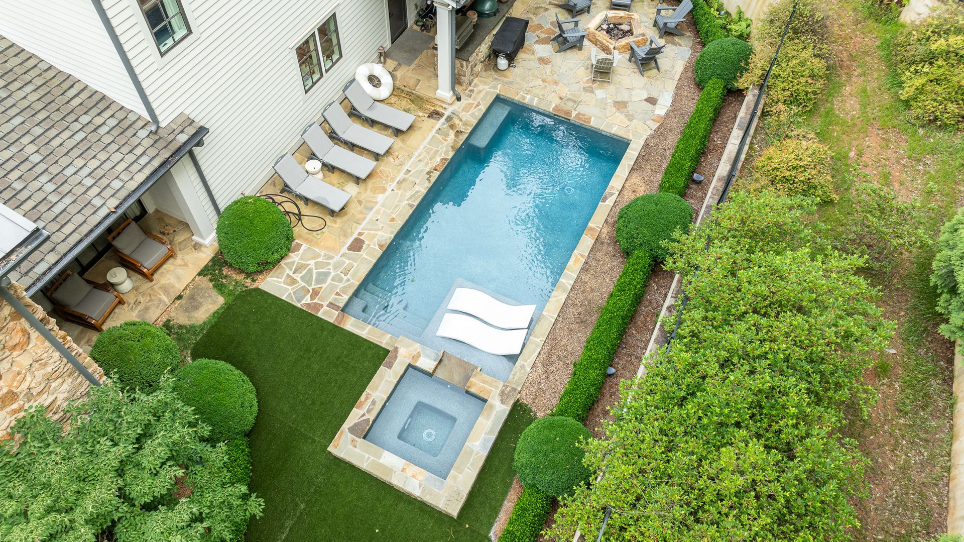 Aerial view of a backyard with a rectangular swimming pool, hot tub, lounge chairs, and green lawn.