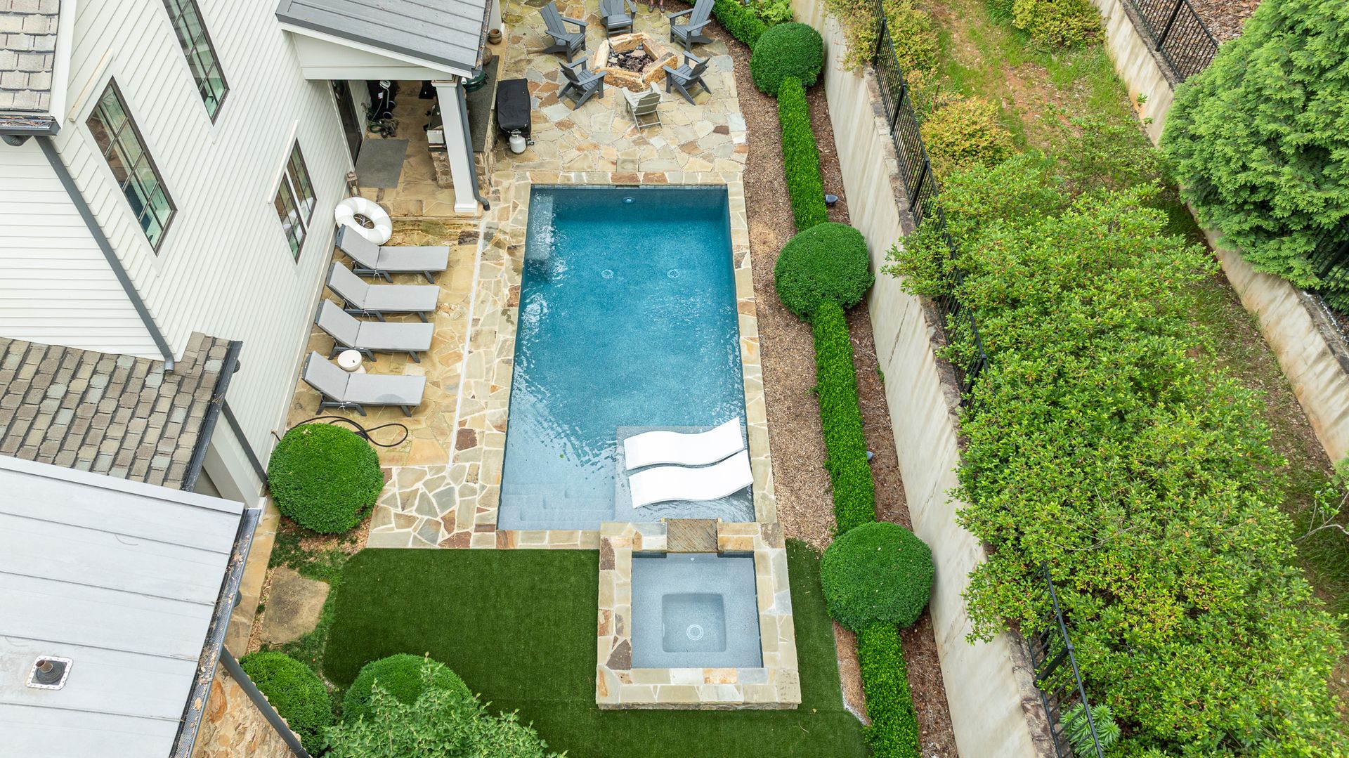 Aerial view of a backyard pool with lounge chairs, hot tub, patio seating, and green landscaping