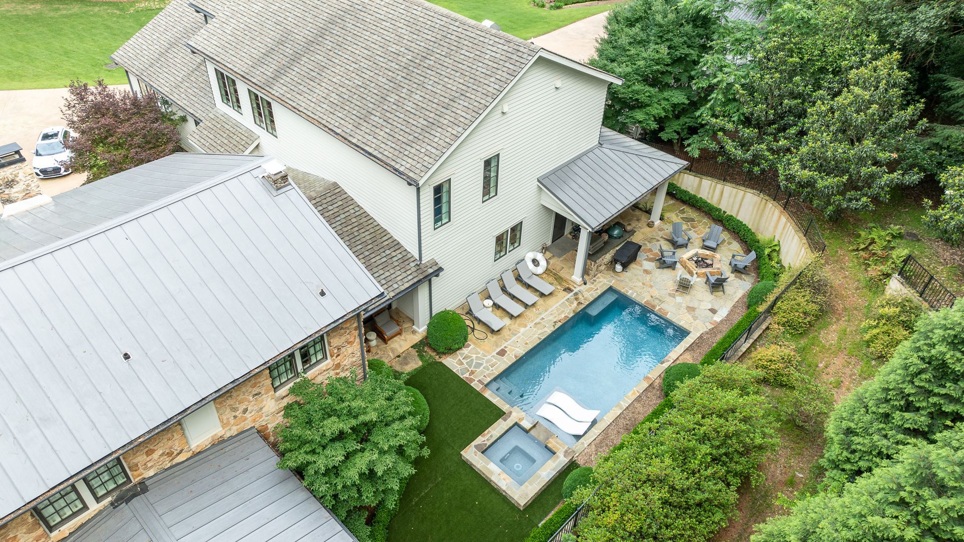 Aerial view of a house with a backyard pool, patio seating, and dense green landscaping