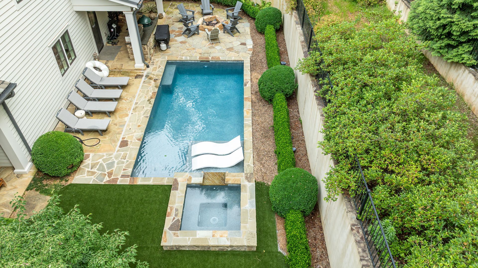 Aerial view of a backyard with a rectangular pool, hot tub, lounge chairs, and trimmed landscaping