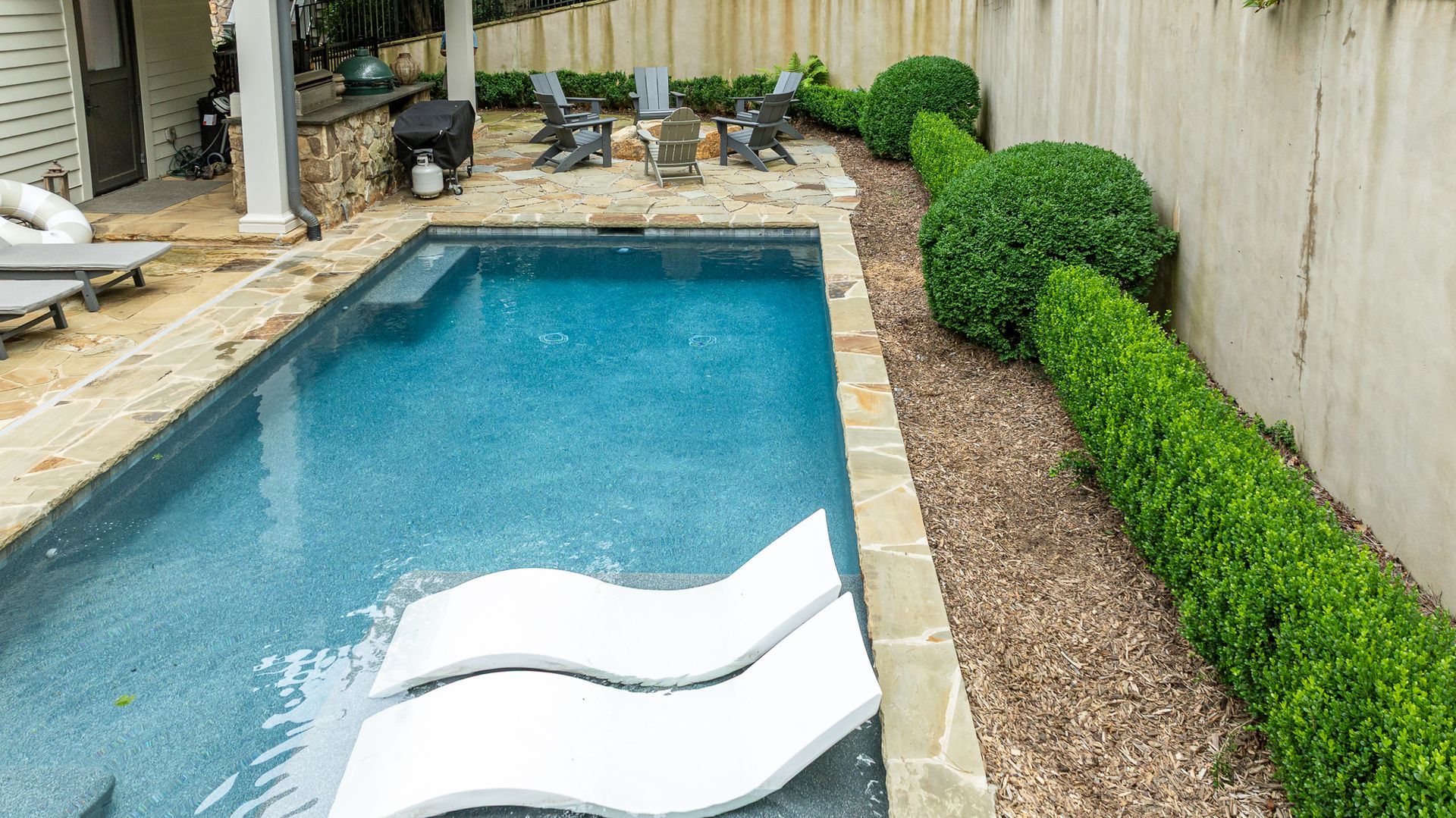 Narrow backyard pool with blue water and two white lounge chairs beside trimmed hedges