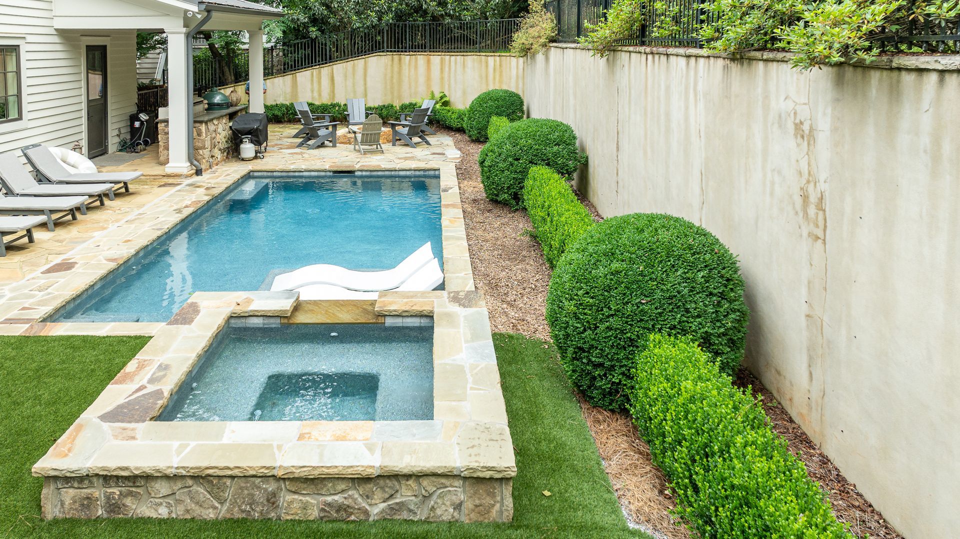 Backyard with a swimming pool and spa, stone patio, trimmed hedges, and a wooden privacy fence