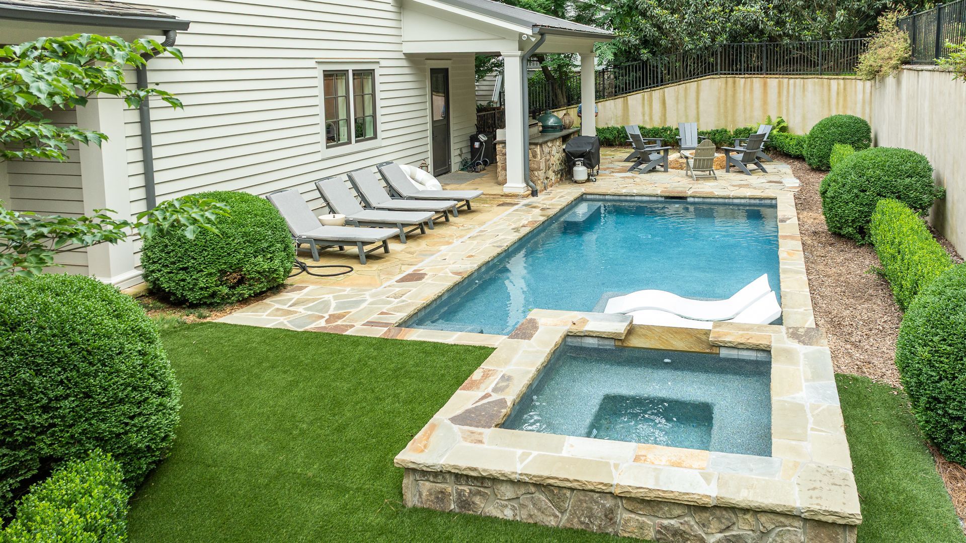 Backyard with a swimming pool, spa, lounge chairs, and manicured greenery beside a house.