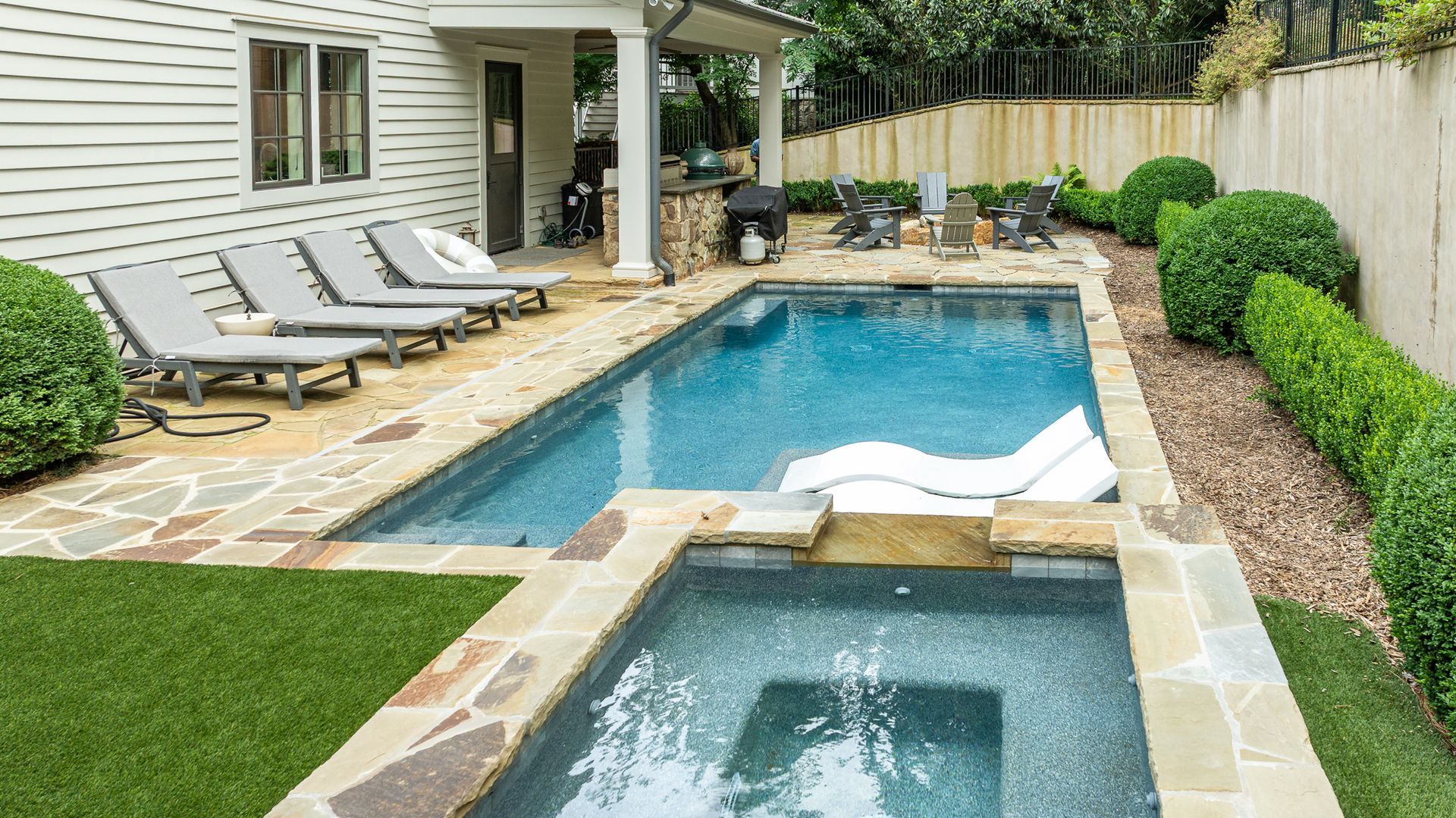 Backyard with a swimming pool, hot tub, lounge chairs, and neatly trimmed shrubs beside a house