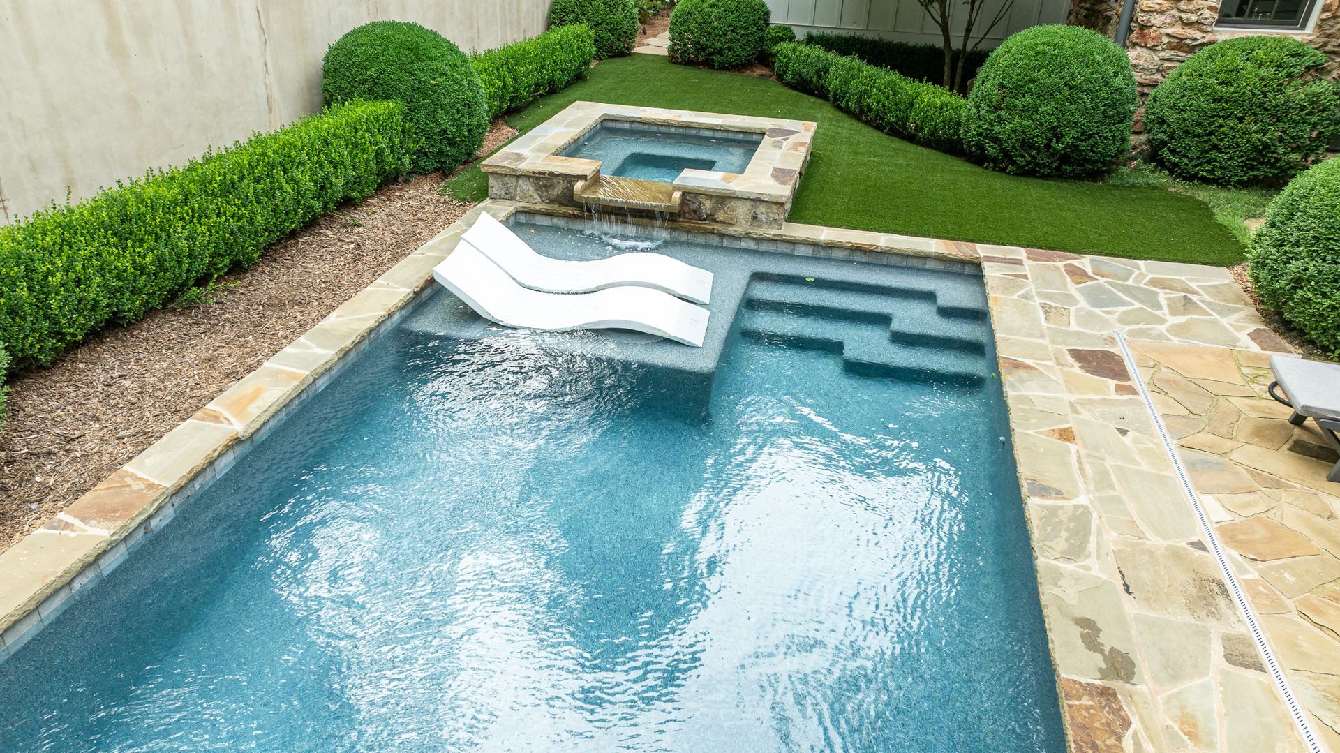 Backyard swimming pool with a built-in spa, white slide, and stone patio surrounded by hedges