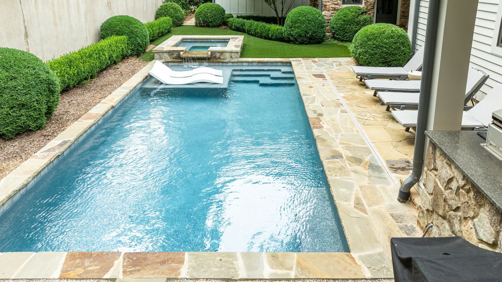 Backyard swimming pool with stone deck, white lounge chairs, and trimmed green hedges