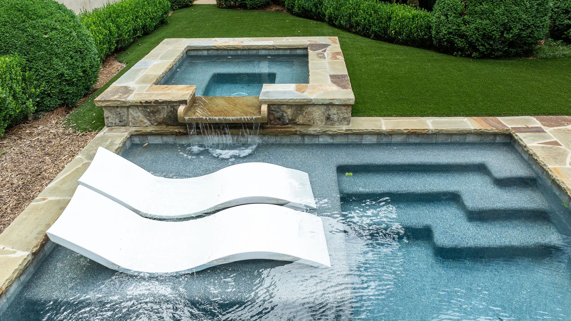 Modern backyard pool with a white slide leading into a blue swimming pool and a raised spa behind it