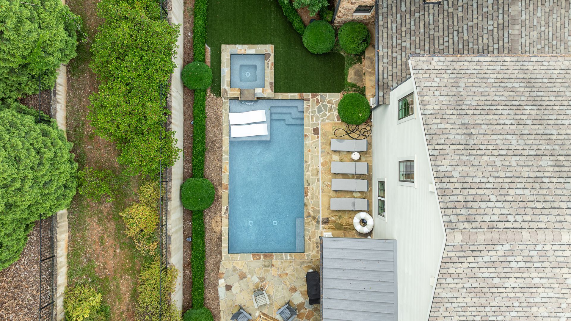 Aerial view of a backyard with a rectangular pool, hot tub, patio seating, and surrounding greenery.