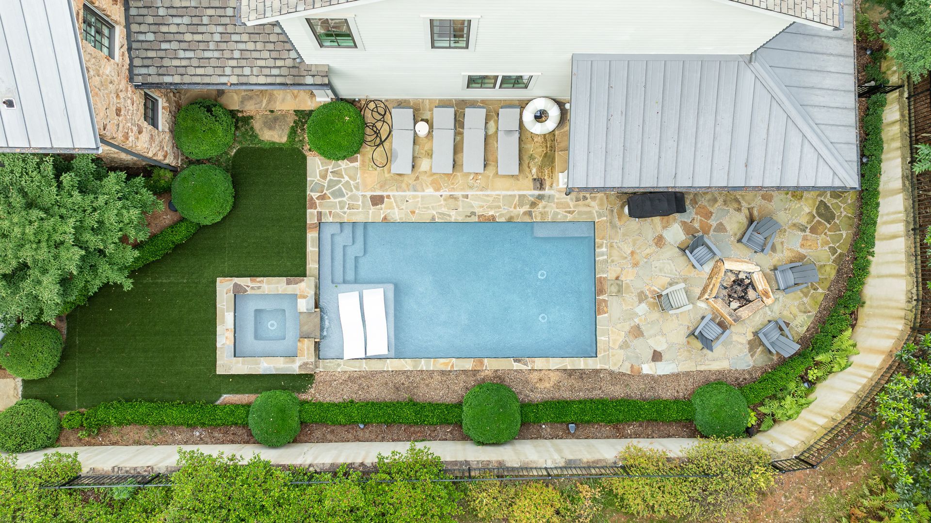 Aerial view of a backyard with a swimming pool, patio seating, lawn, and surrounding hedge.