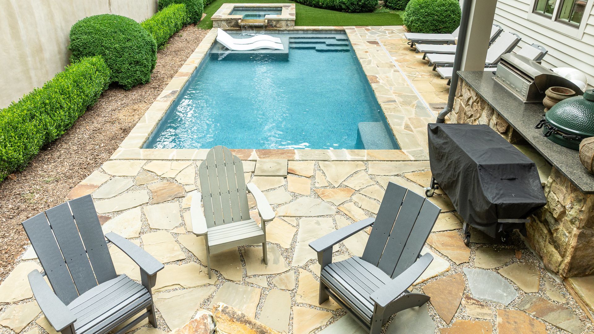 Stone patio with lounge chairs beside a rectangular backyard swimming pool and grill
