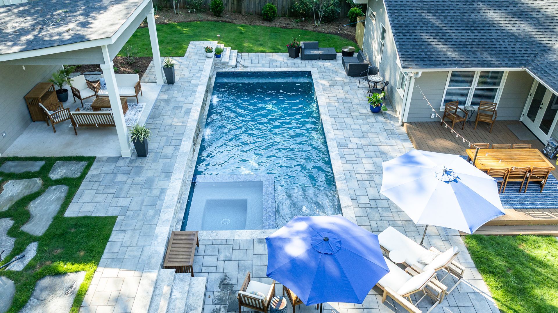 Aerial view of a backyard with a rectangular pool, patio seating, umbrellas, and a covered lounge area.