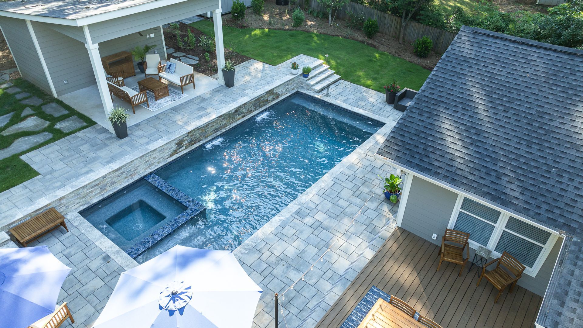 Aerial view of a backyard pool with spa, stone patio, covered seating, and a house deck.