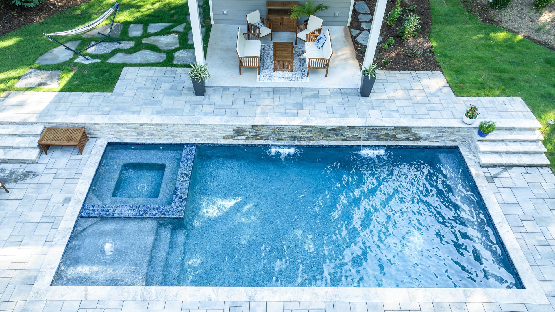 Aerial view of a rectangular backyard pool with stone patio, lounge chair, and covered seating area.