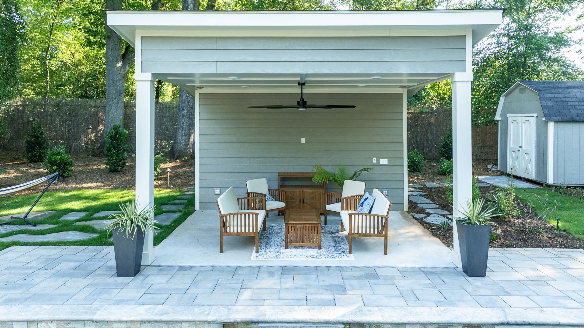 Covered outdoor patio with seating, ceiling fan, and potted plants in a landscaped backyard