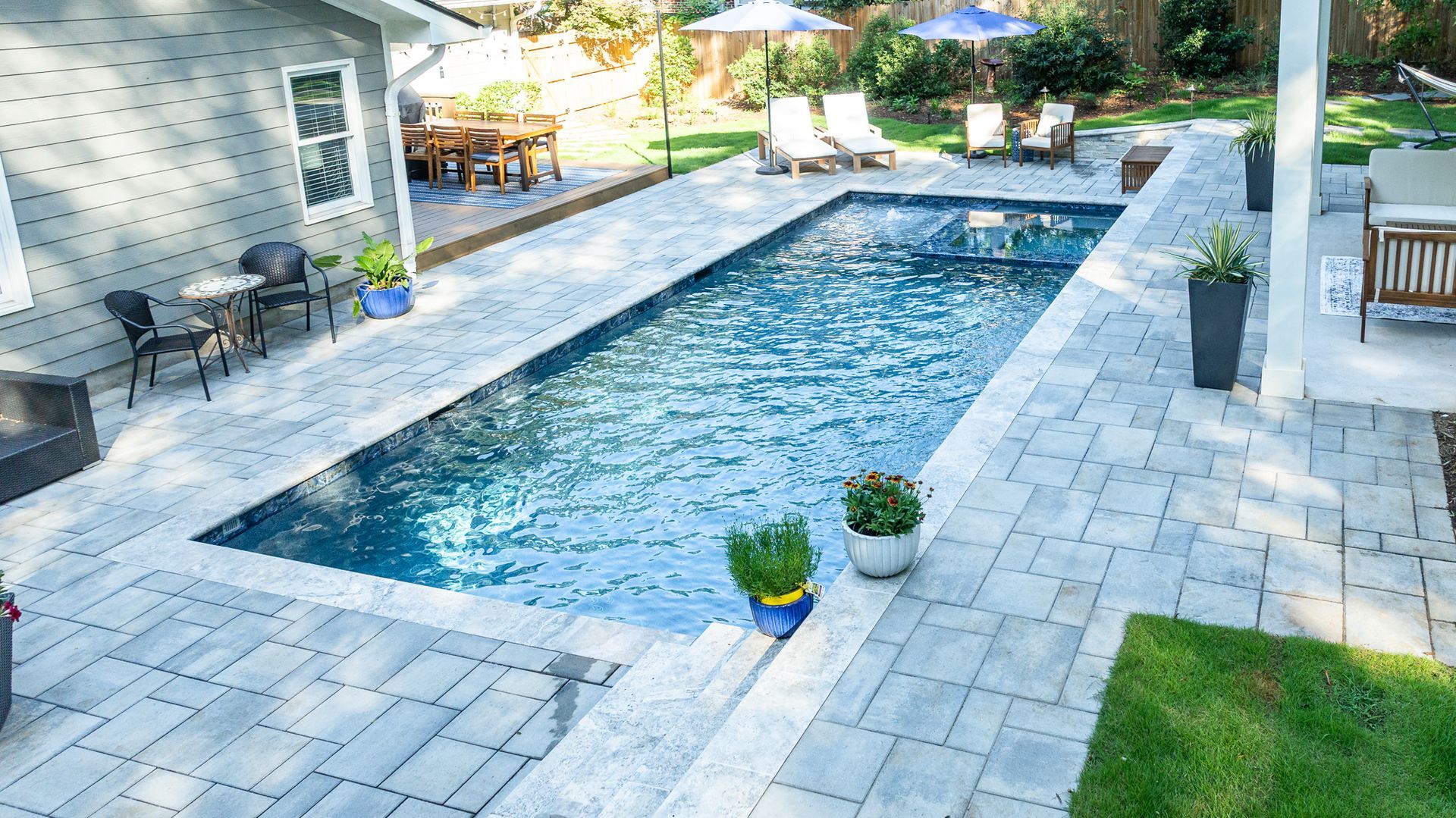 Backyard pool surrounded by light stone patio, lounge chairs, potted plants, and a house exterior