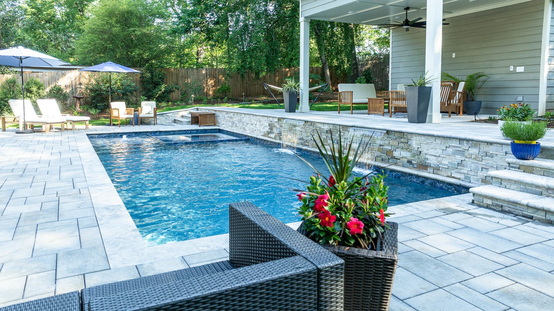 Backyard pool with stone patio, wicker chairs, and potted flowers beside a house