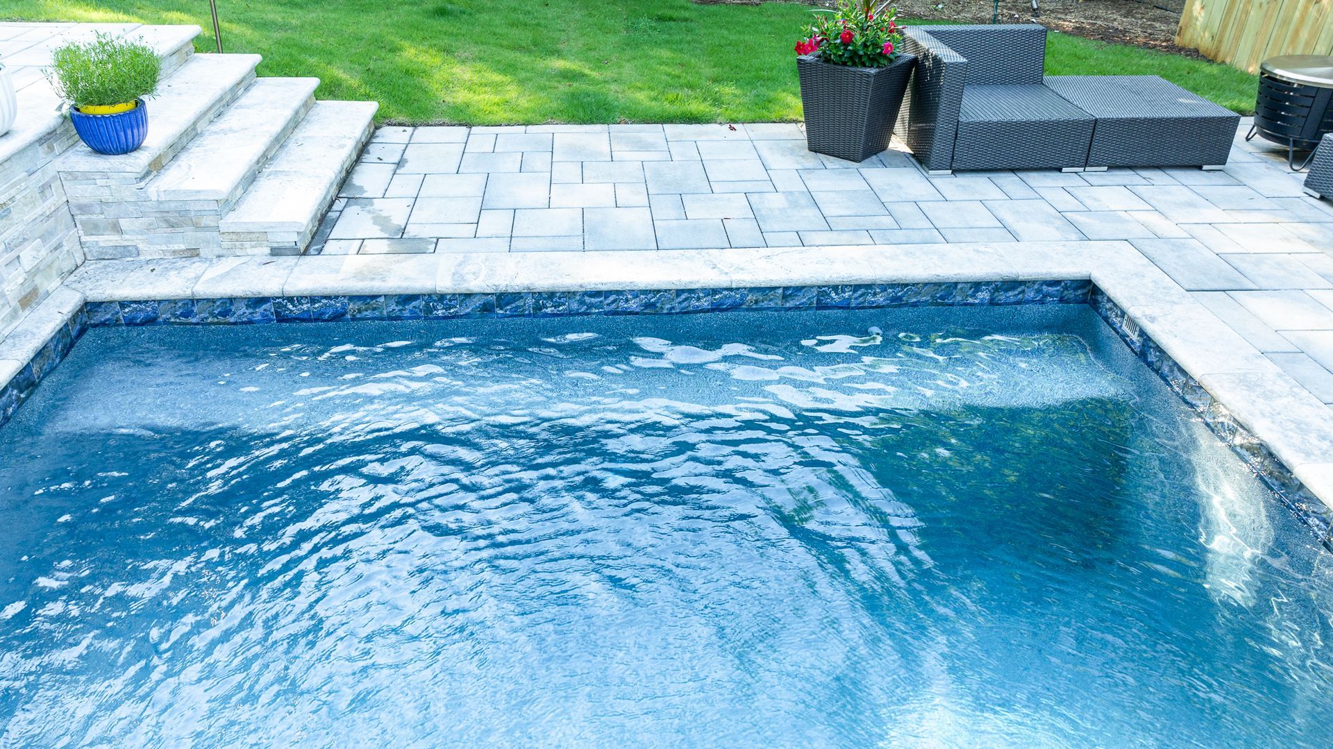 Blue swimming pool beside a stone patio with potted plants and lawn in the background