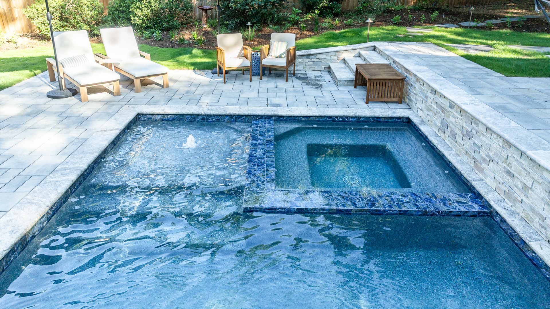 Modern backyard pool with built-in spa, stone deck, lounge chairs, and a small seating area