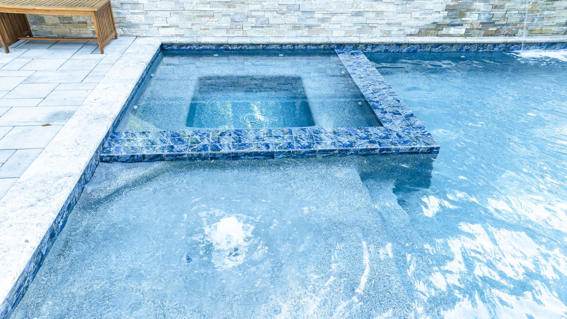 A blue tiled pool with a raised square spa and clear water reflecting sunlight