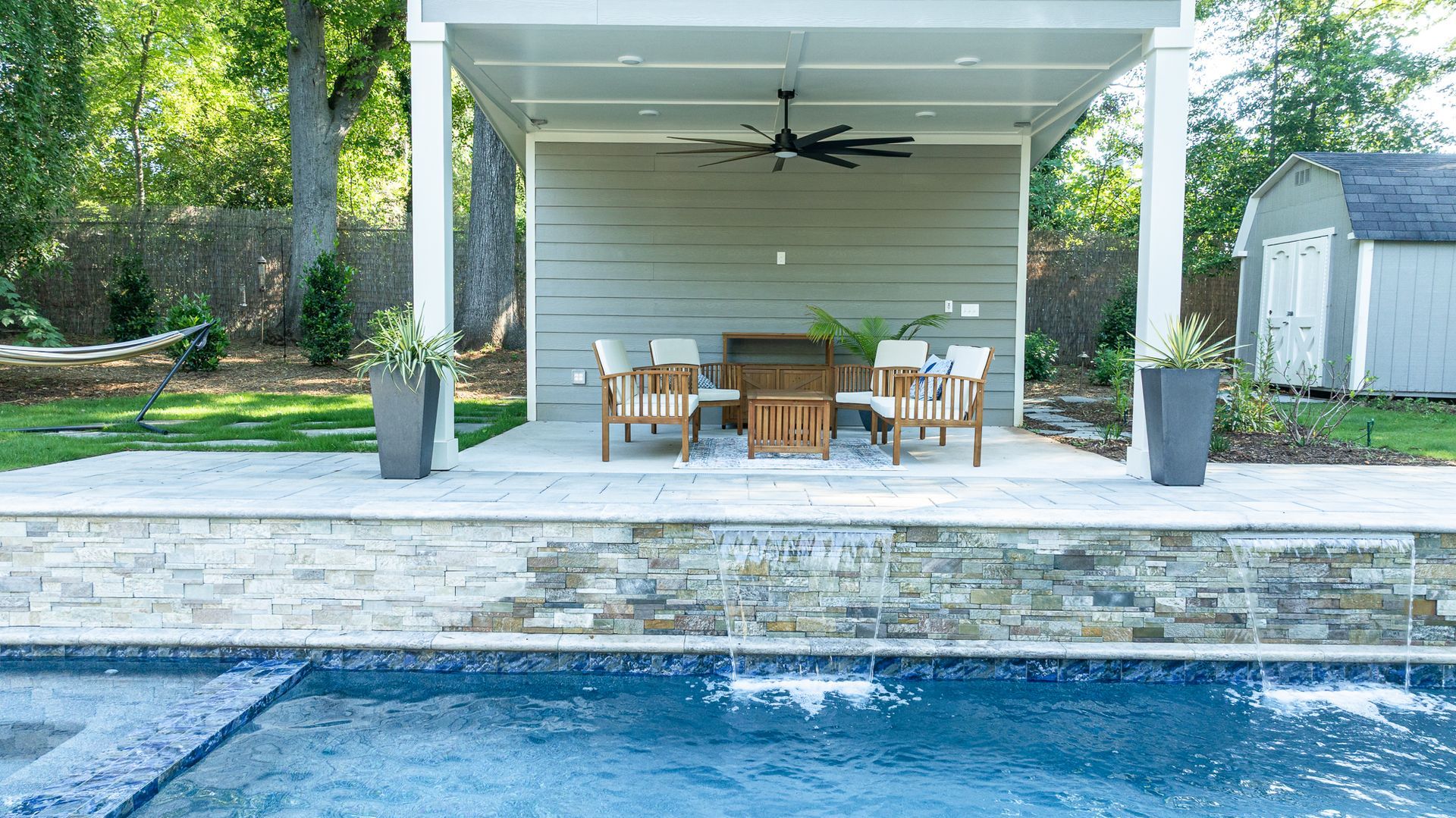 Backyard pool beside a covered patio with outdoor dining table, chairs, and ceiling fan
