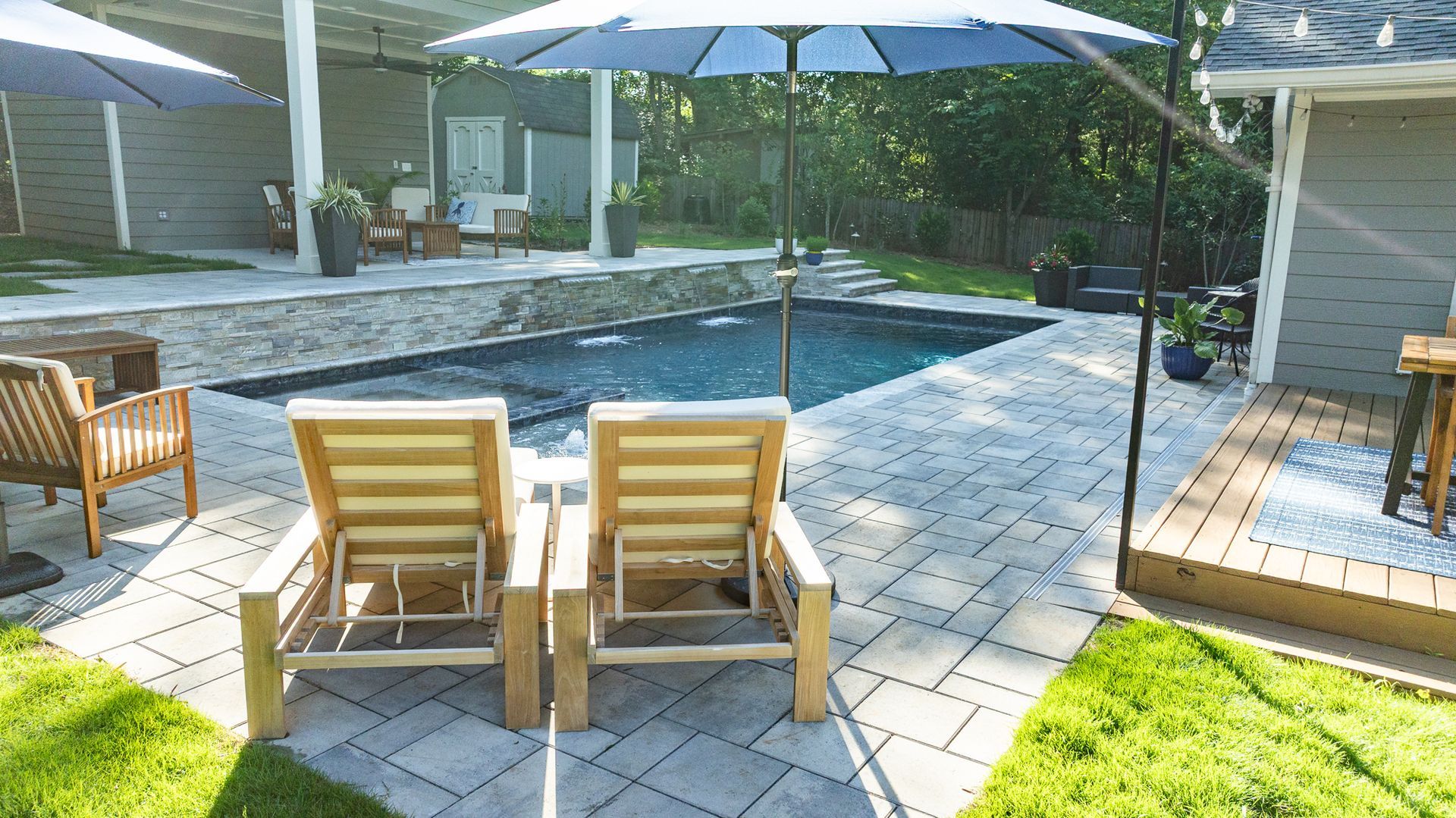 Backyard patio with two lounge chairs, umbrella, pool, and wooden decks in bright sunlight