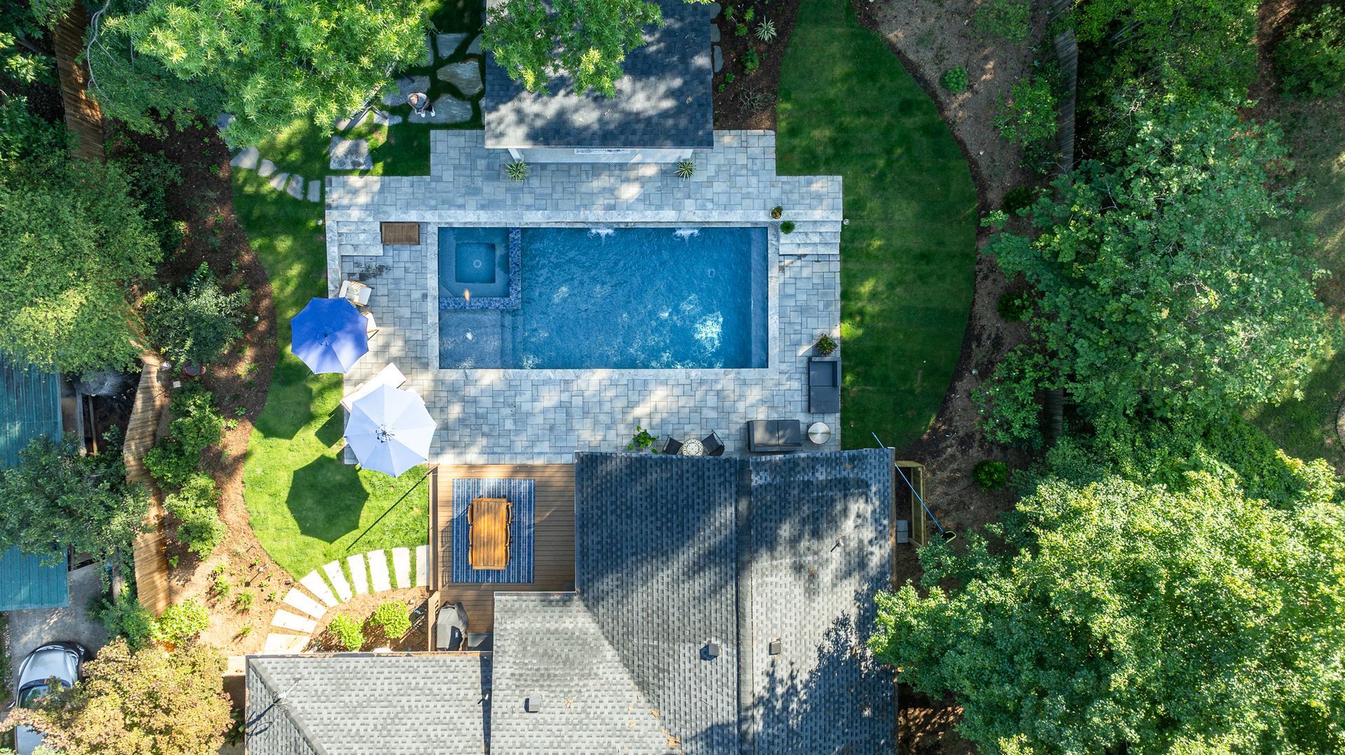 Aerial view of a backyard with a rectangular blue pool, patio seating, and lush surrounding trees.