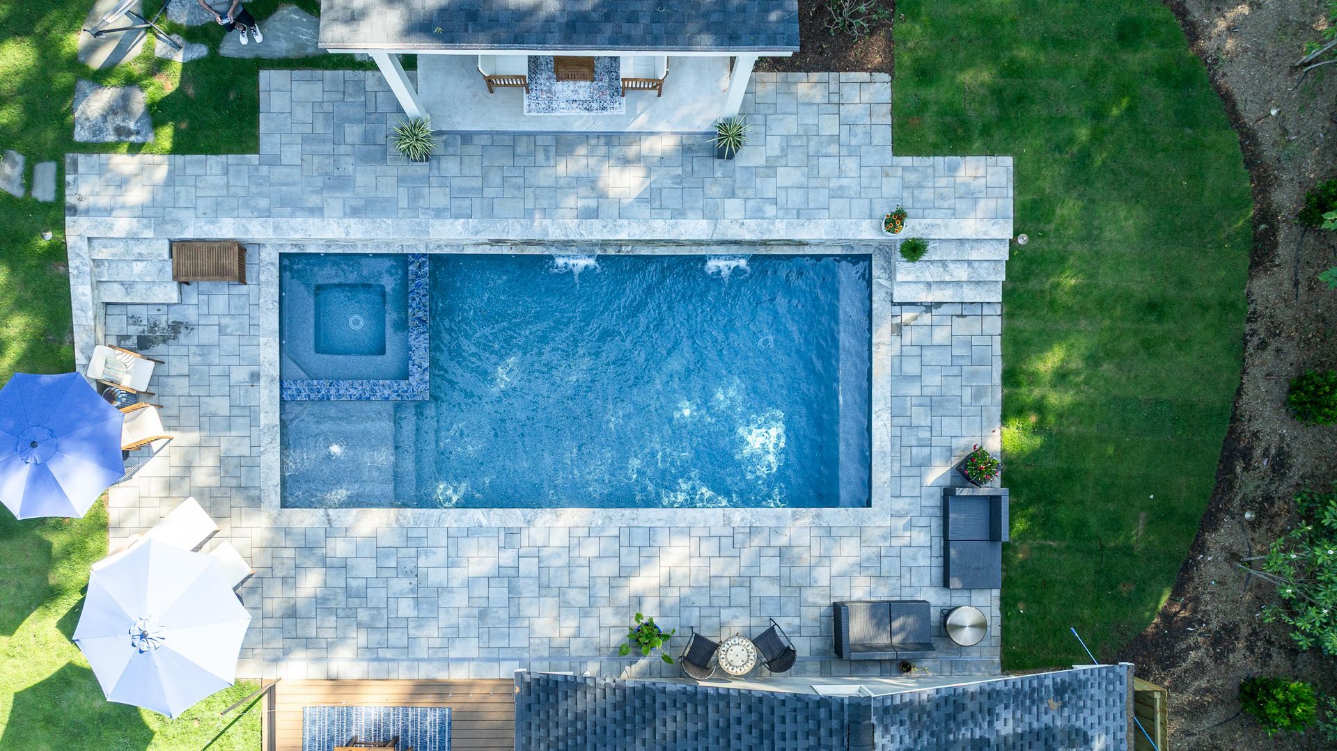 Aerial view of a backyard pool with stone patio, lounge chairs, and surrounding green lawn.