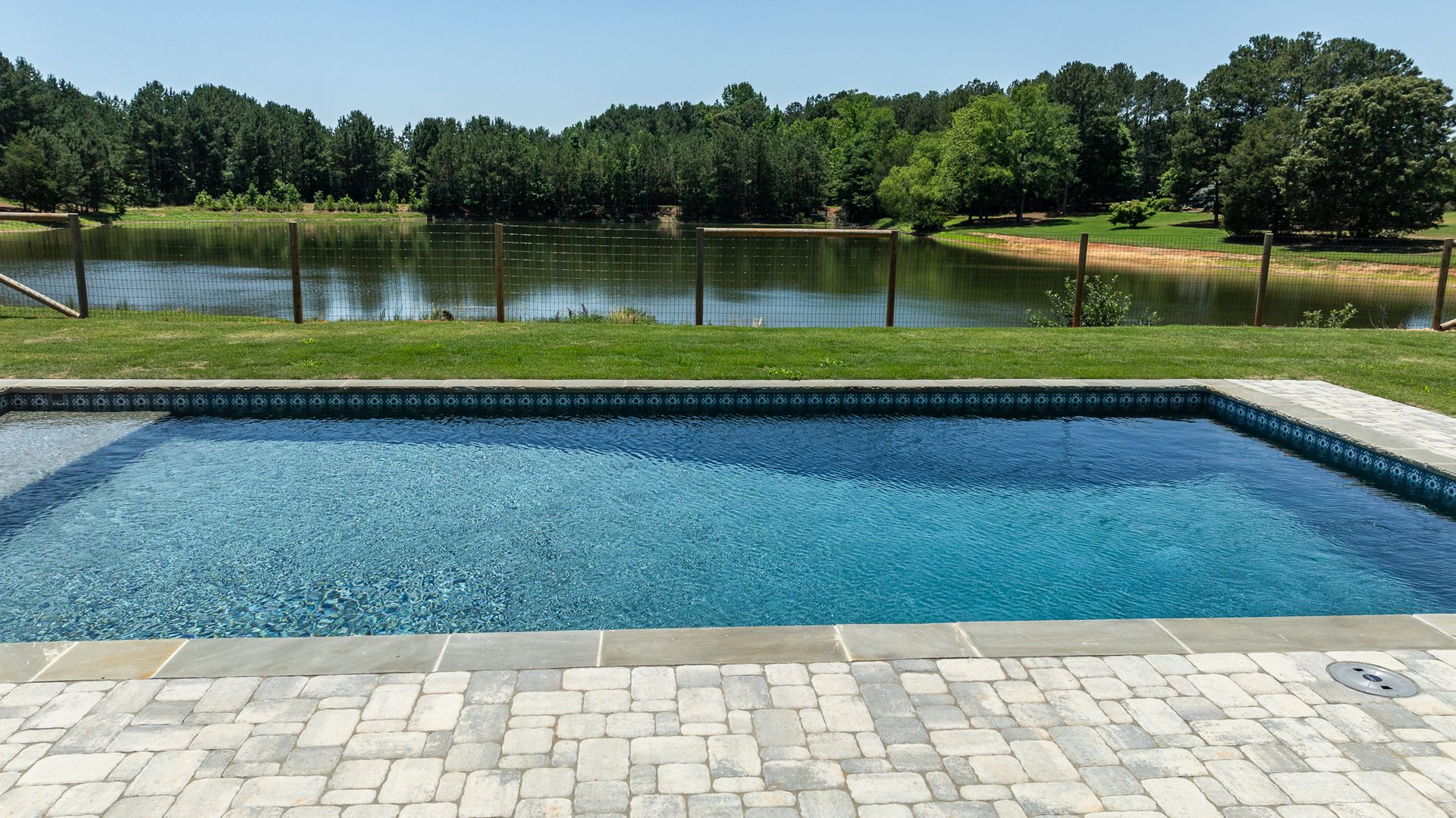 Rectangular backyard pool with blue water beside a lake and trees under a clear sky