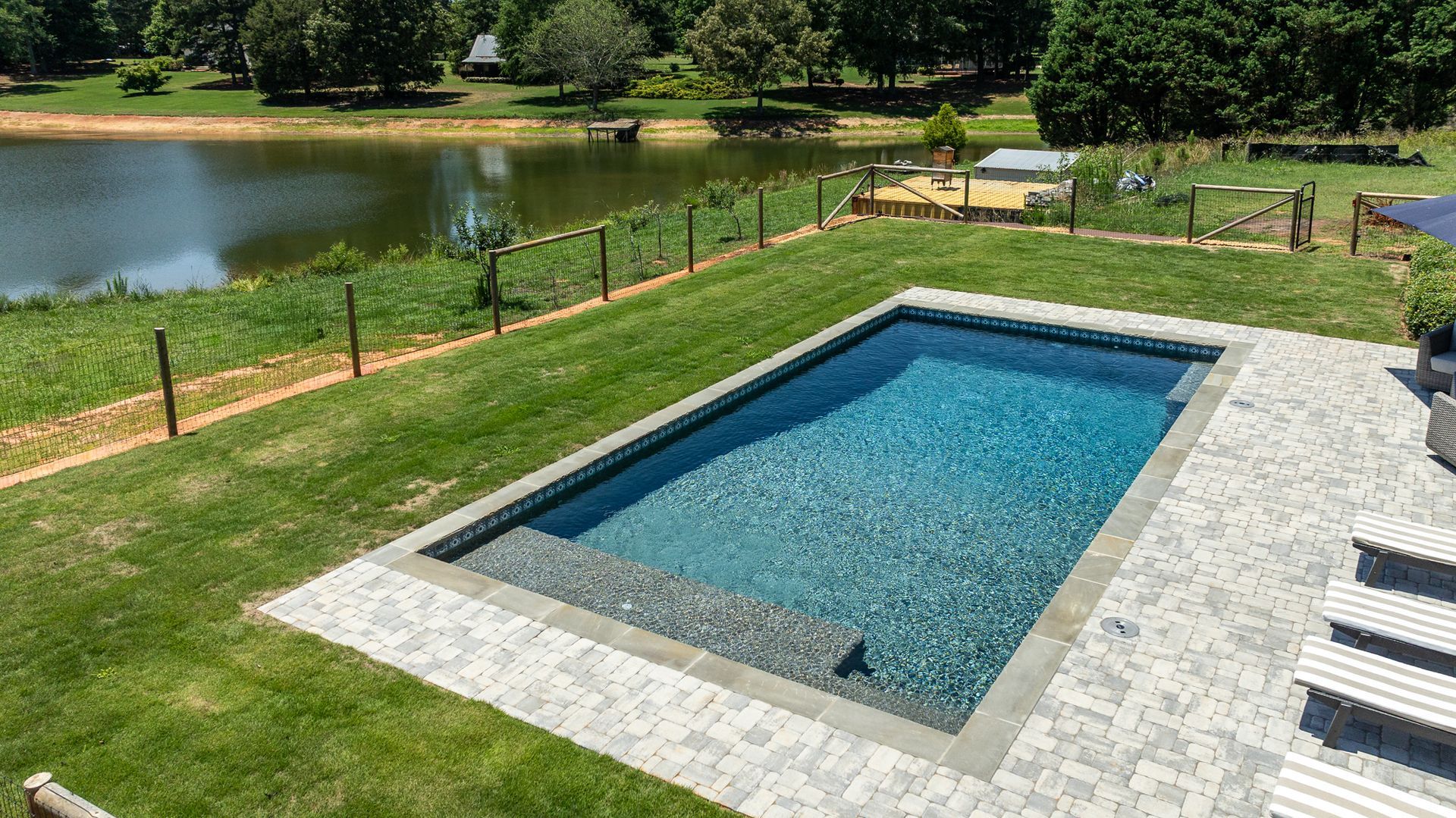 Rectangular blue pool beside a grassy yard and lake, with stone patio and black fence surrounding it