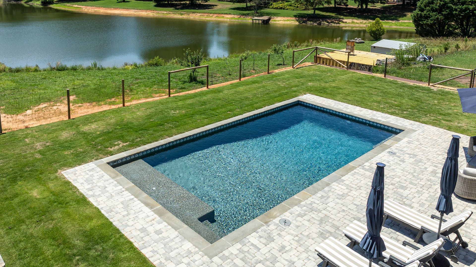 Rectangular blue swimming pool beside a grassy backyard and lake, with patio chairs and umbrellas nearby