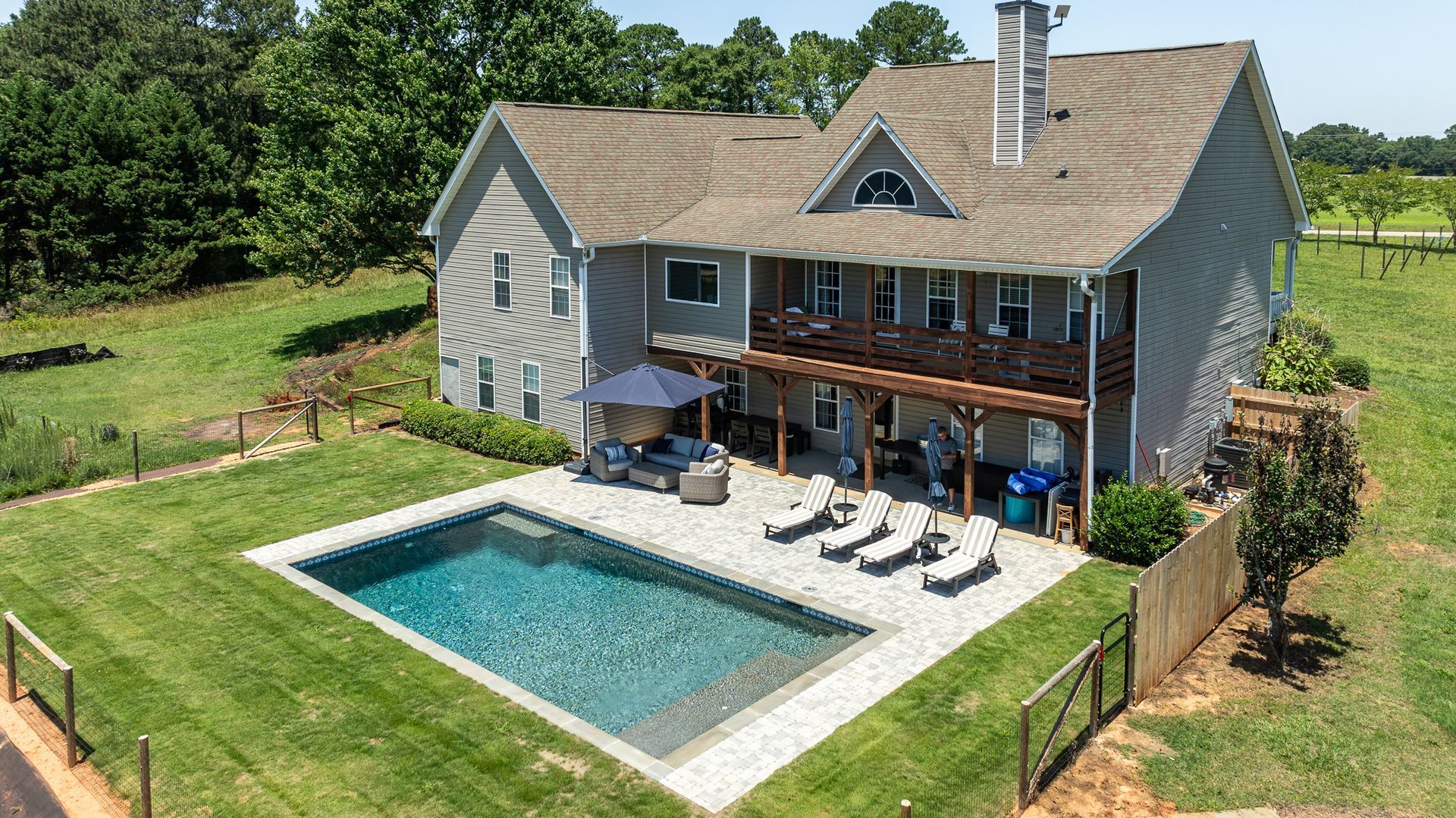 Backyard pool beside a gray two-story house with a wooden deck and patio chairs