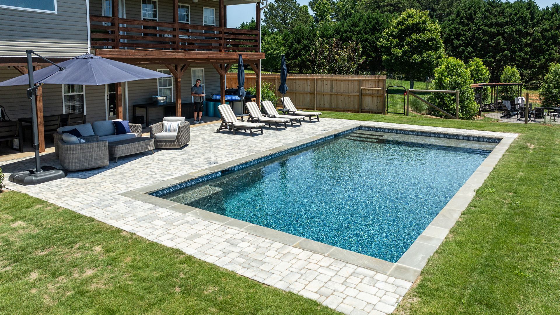 Backyard pool with stone deck, lounge chairs, umbrella, and patio seating beside a house