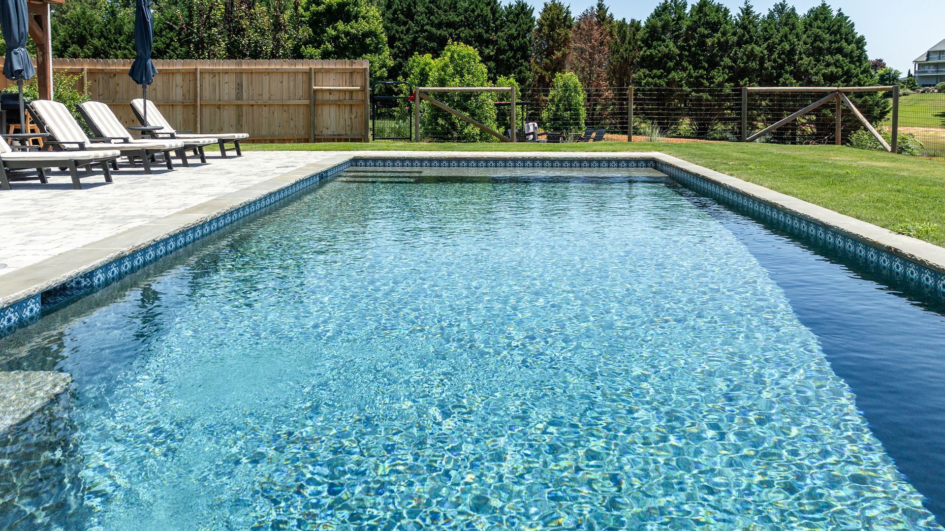Backyard swimming pool with clear blue water, lounge chairs, and green lawn under sunny skies