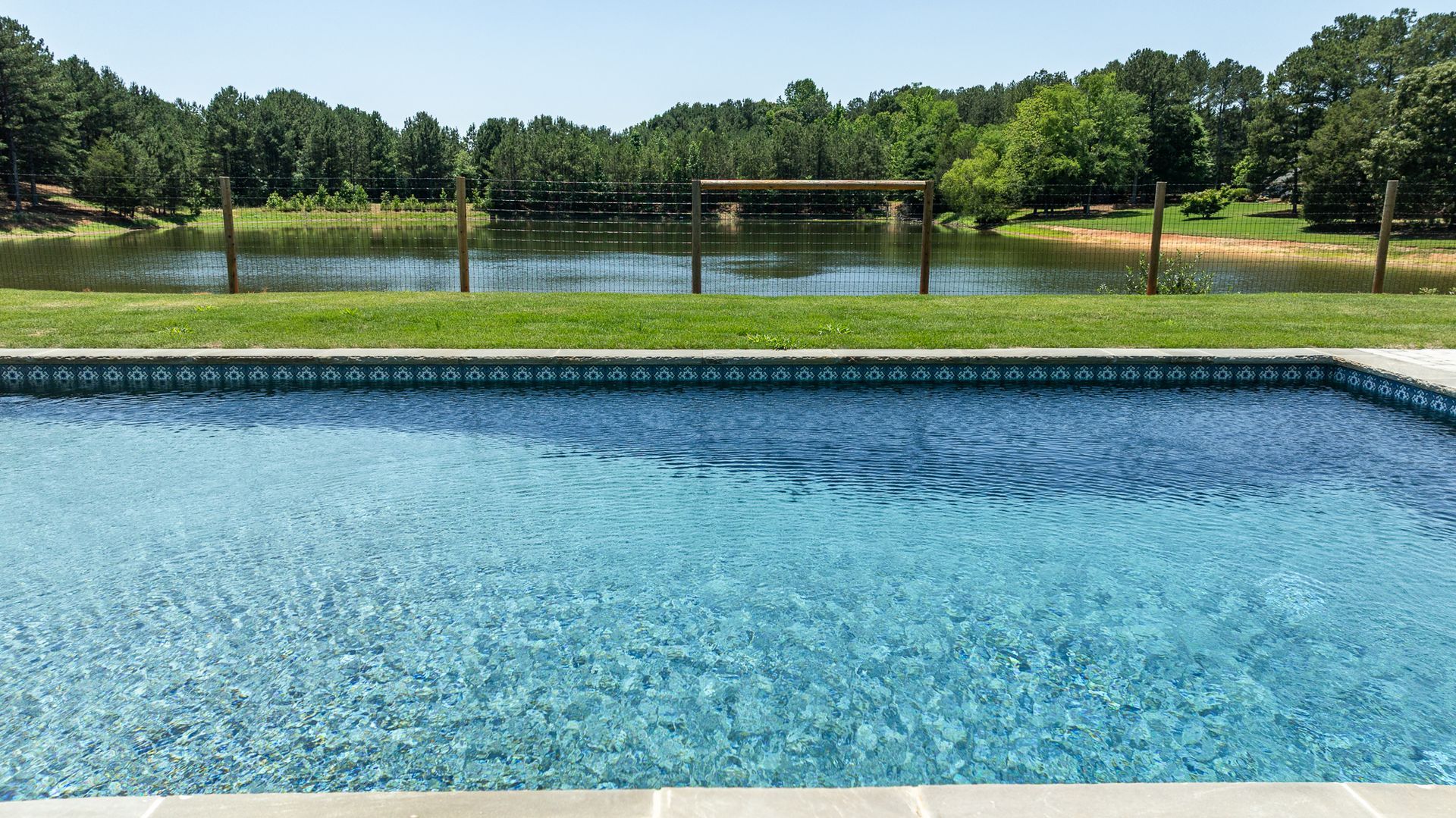 Blue swimming pool in the foreground with a lake, trees, and a sunny park in the background