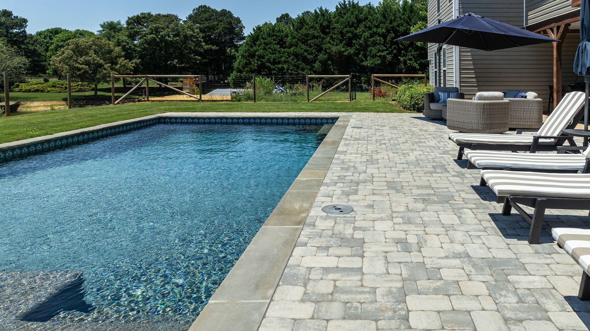 Backyard swimming pool beside a stone patio with lounge chairs and trees in the background