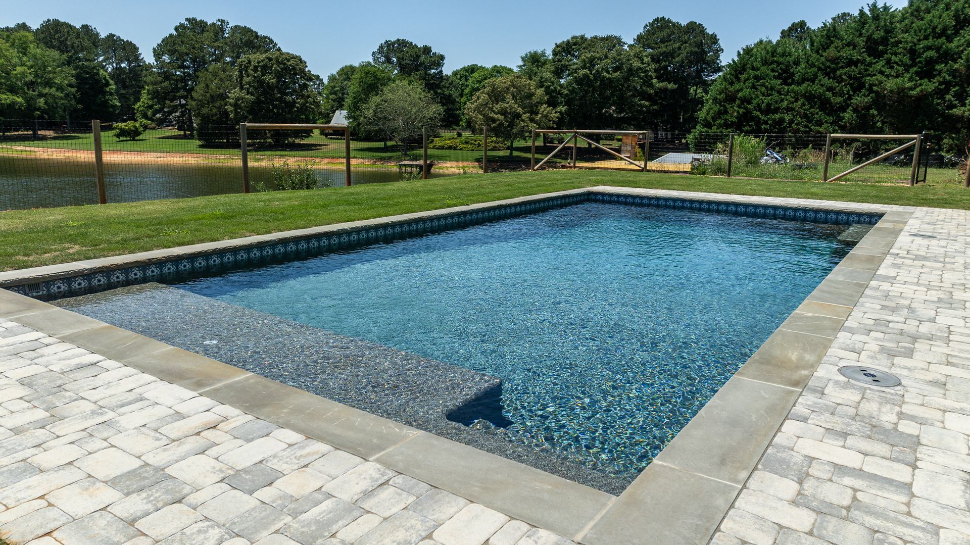 Rectangular outdoor swimming pool with blue water, surrounded by light stone paving and trees in the background
