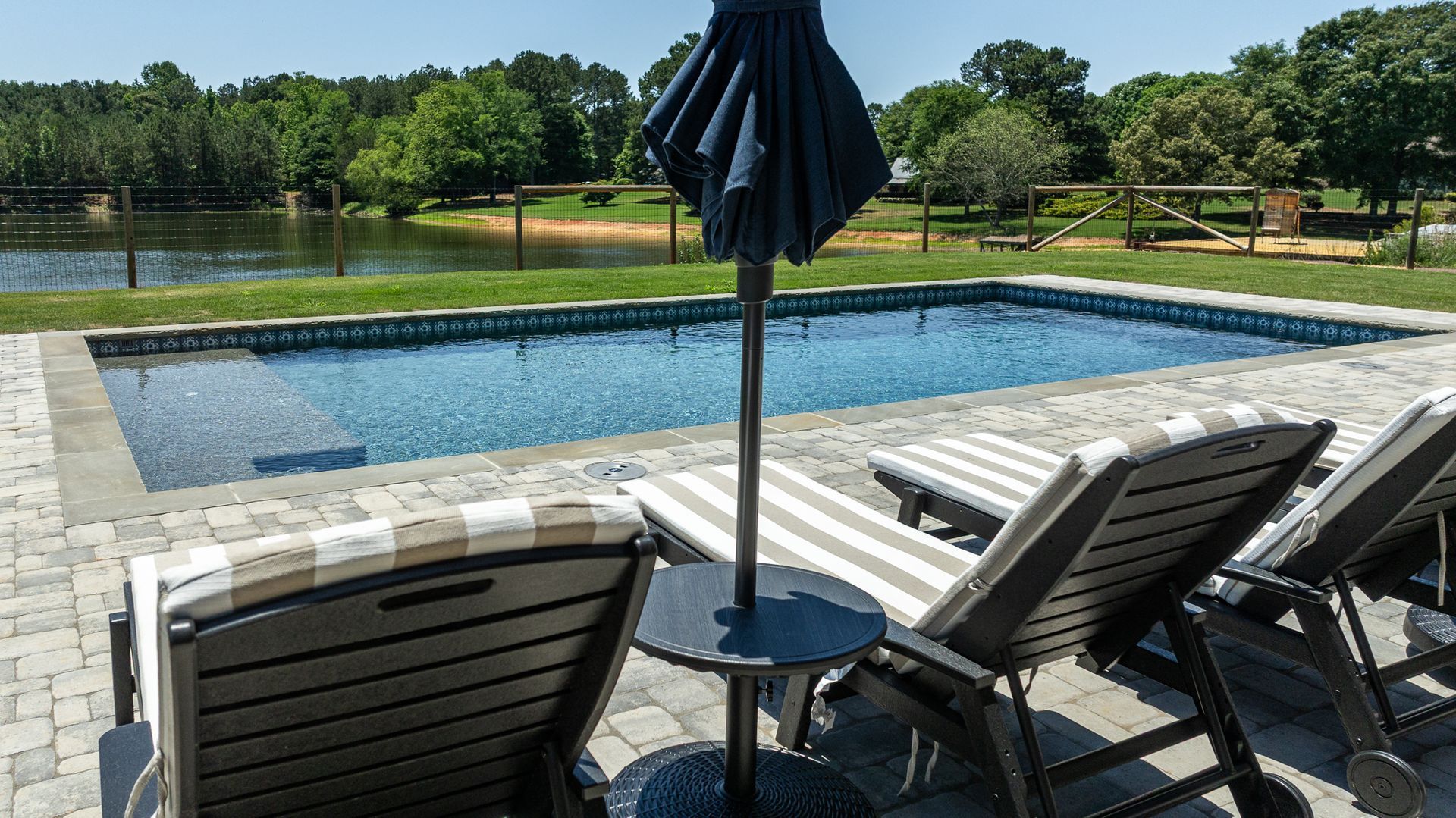 Patio chairs and closed umbrella facing a backyard pool with trees and water beyond