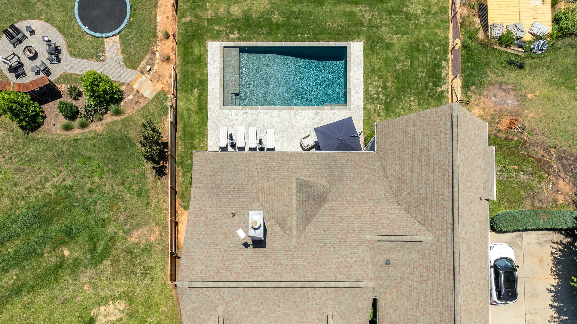 Aerial view of a house with a backyard pool, patio chairs, lawn, and a parked car nearby.