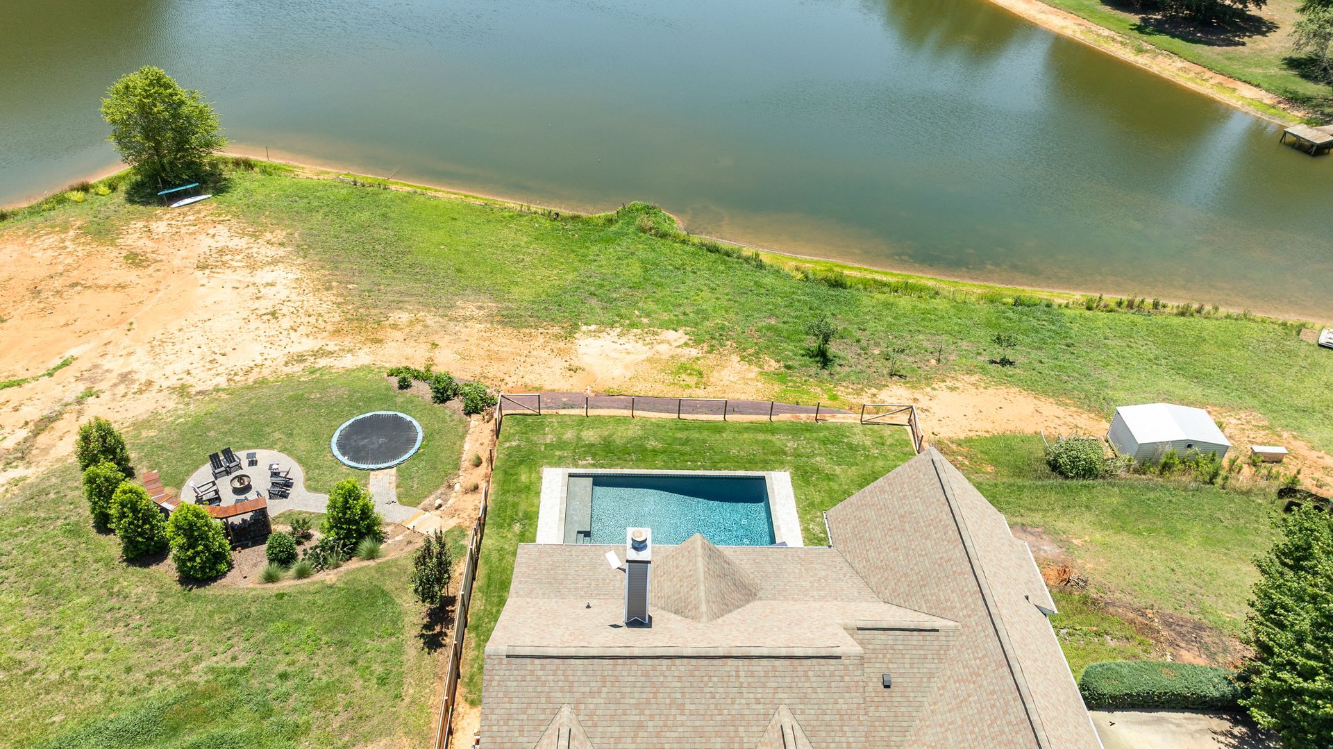 Aerial view of a house by a pond with a patio, trampoline, and grassy yard