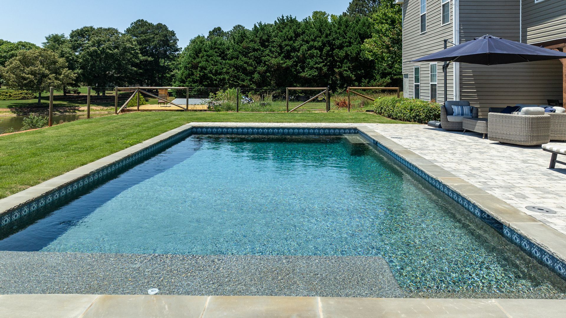 Rectangular backyard swimming pool with clear blue water beside a patio and shaded outdoor seating area