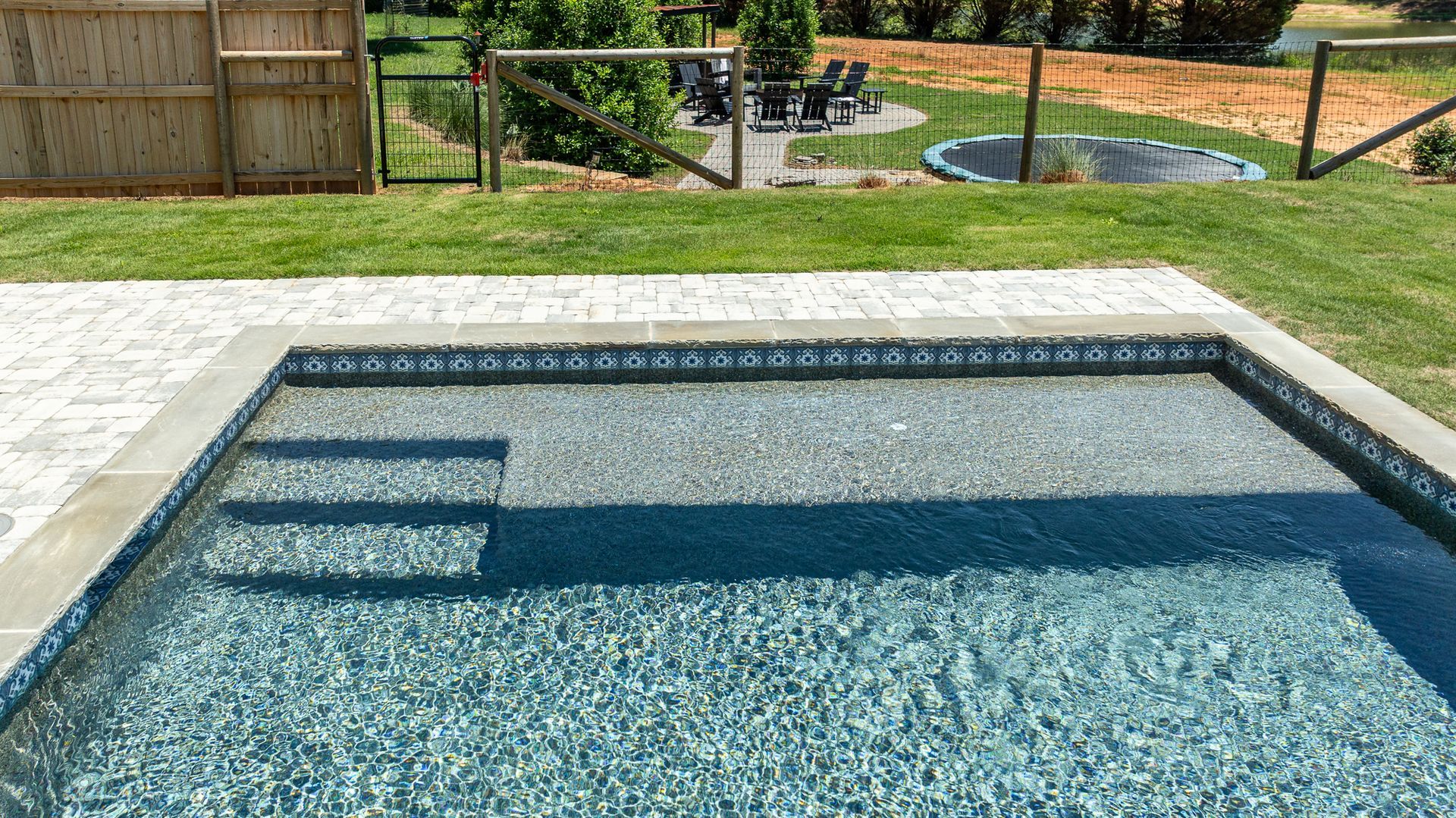Backyard swimming pool with clear blue water, steps, and stone coping, overlooking a grassy yard.