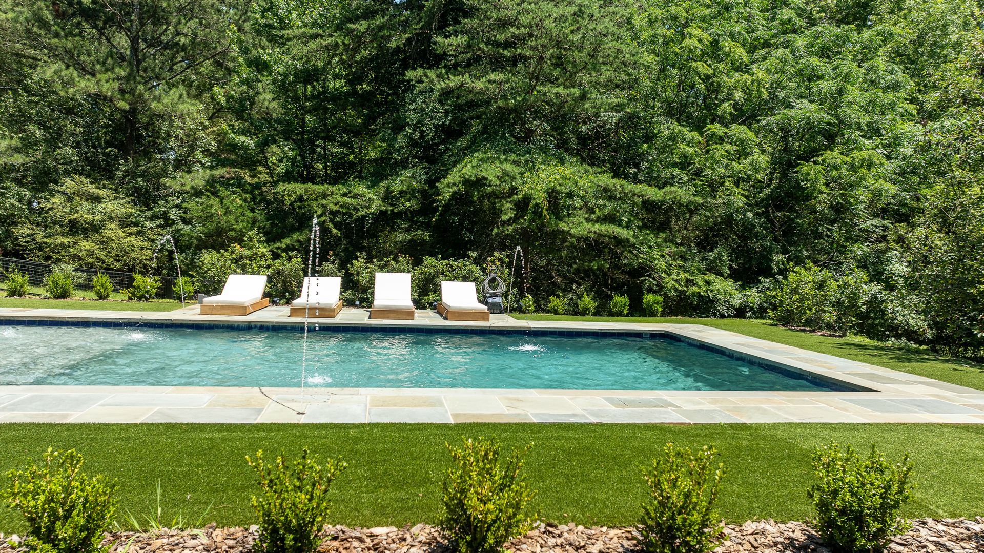 Rectangular backyard pool with lounge chairs, bordered by lush green trees and lawn