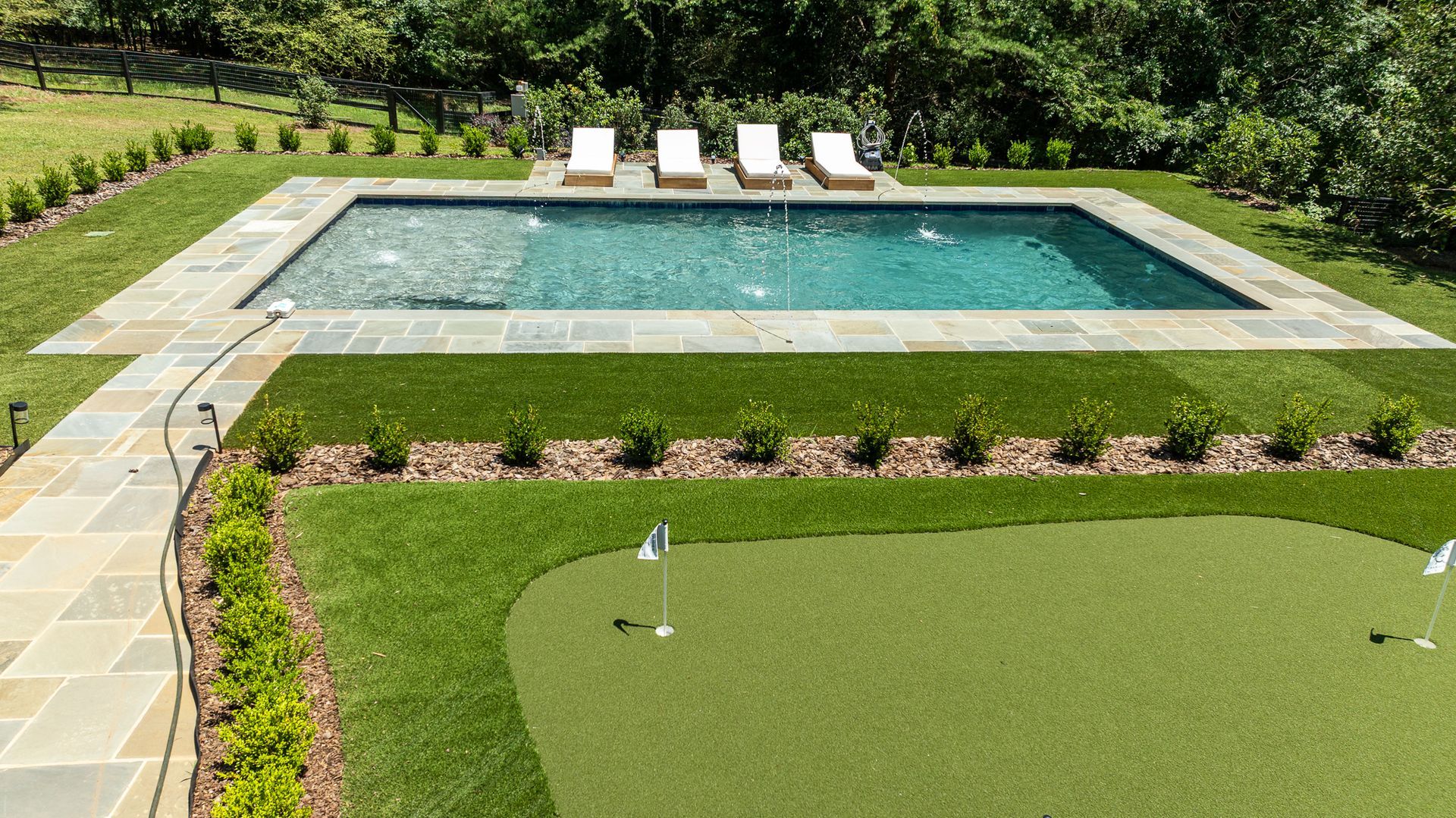 Backyard pool with stone patio, lounge chairs, and a green lawn bordered by shrubs