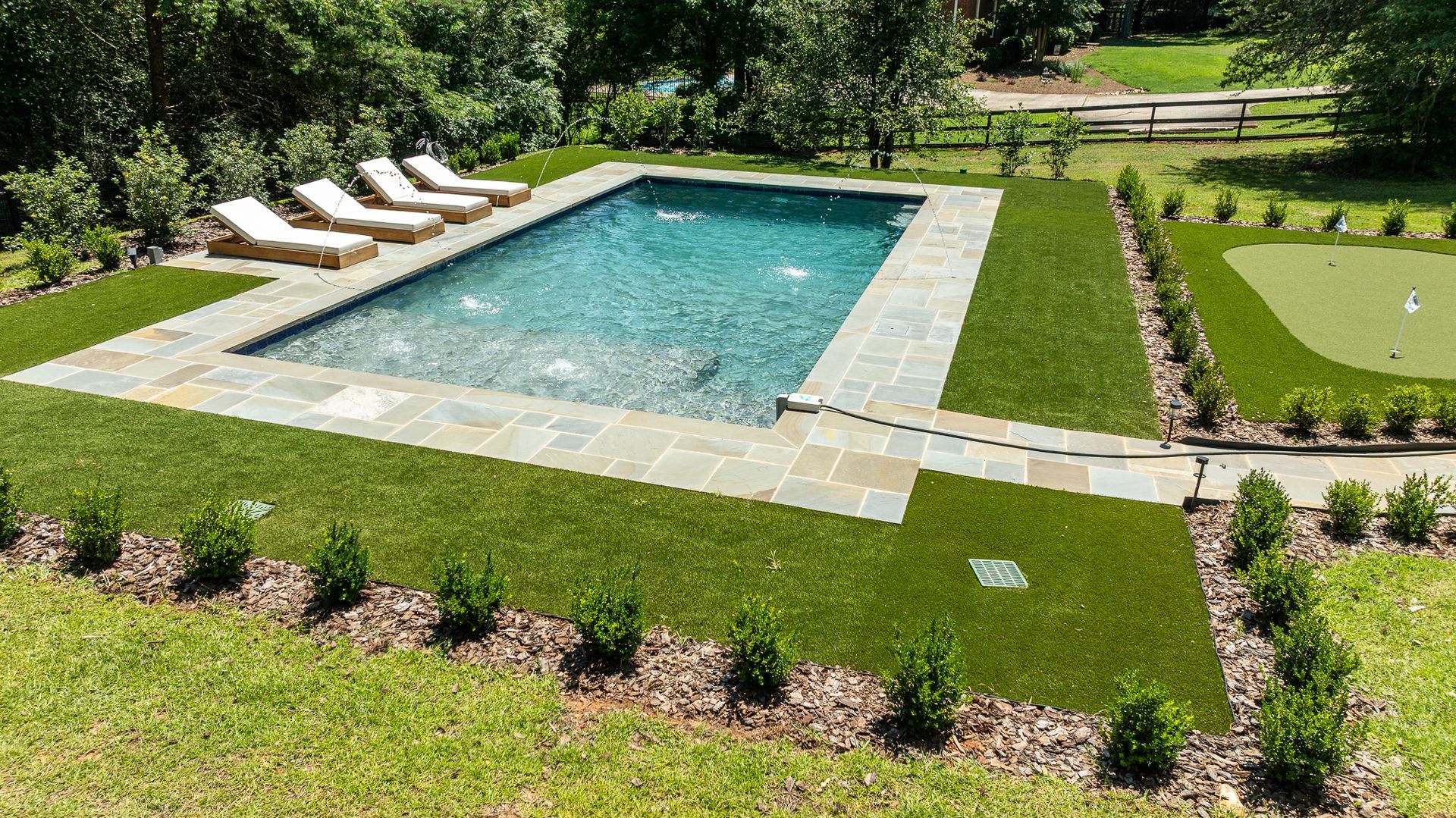 Rectangular backyard pool with four lounge chairs on a stone patio, surrounded by manicured lawn and garden beds